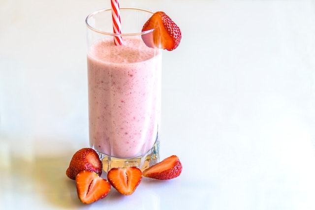 Vários copos de smoothies coloridos alinhados em uma mesa de madeira, com uma variedade de frutas frescas como morangos, bananas e kiwis ao fundo.